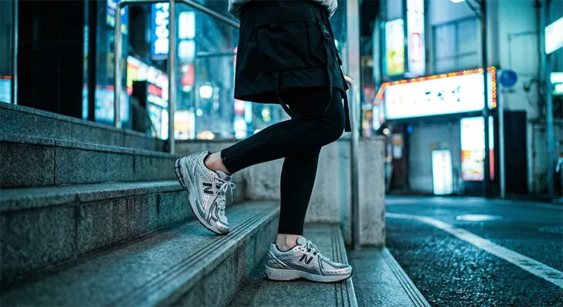 밤 네온사인을 반사하는 실버 뉴발란스 1906R 운동화 착용 샷. 테크웨어 미니스커트와 레깅스를 입은 여성이 도시 계단을 내려온다.