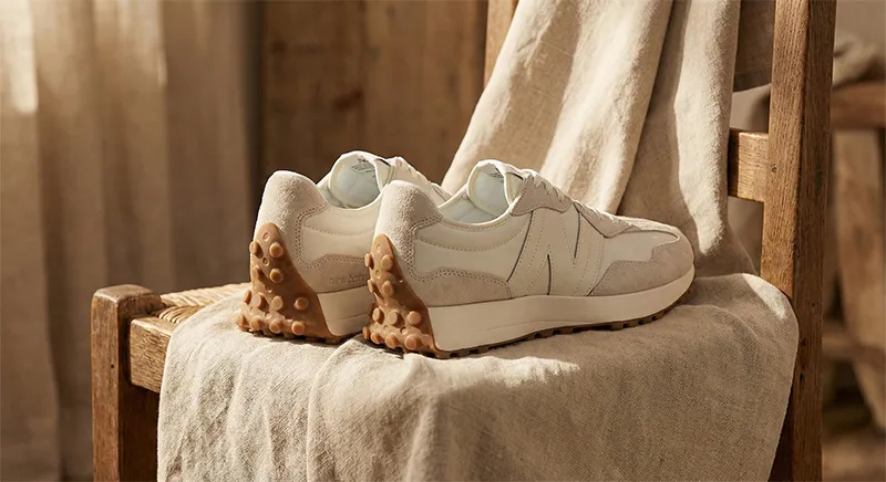 따뜻한 햇살 아래 린넨 천이 덮인 나무 의자 위 뉴발란스 327 씨 솔트 운동화. 뒤축 스터드 밑창이 돋보이는 정물 사진.