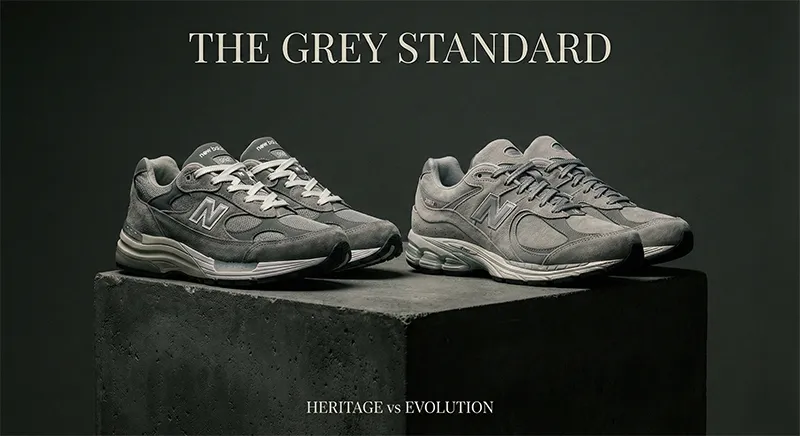 콘크리트 큐브 위에 뉴발란스 992와 2002R 그레이 스니커즈가 나란히 놓여 있고, 상단에 'THE GREY STANDARD' 텍스트가 있는 흑백 정물 사진입니다.
